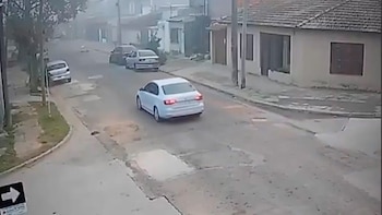 Vista aérea de una calle con tres automóviles, casas bajas a los lados y árboles en un día nublado. La calle presenta baches y un cartel de dirección