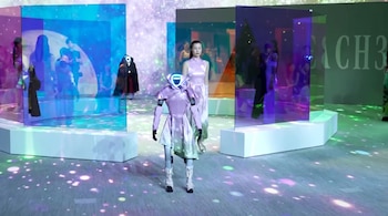 Una mujer vestida de morado y un robot humanoide a su lado caminan sobre un suelo iluminado con destellos, rodeados de paneles translúcidos que muestran figuras.