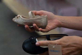 Dos manos sostienen dos zapatos infantiles: uno dorado con brillo y una correa beige, otro negro con lazo. Se ve una etiqueta de cartón