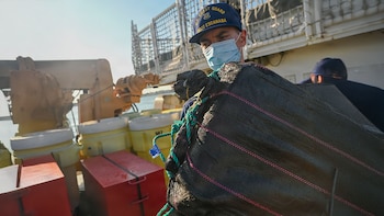 Miembro de la Guardia Costera de EE.UU. con mascarilla y gorra "USCGC Escanaba" manipula un gran fardo oscuro en la cubierta de un barco, con contenedores visibles