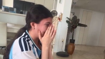 Una mujer, vista de perfil, cubriendo su cara con las manos. Lleva una camiseta de rayas blancas y celestes. El fondo incluye una cocina, una planta y una pared