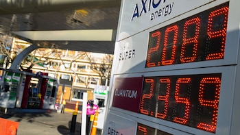 Primer plano de un cartel electrónico de precios de Axion Energy con números rojos, mostrando los precios de combustibles Super y Quantium. Al fondo, surtidores y un edificio