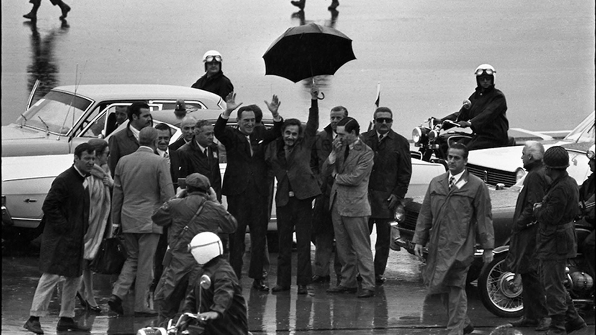 Secuencia de imágenes del regreso, luego de 18 años de exilio, de Juan Domingo Perón a Argentina en el que José Ignacio Rucci sostiene el paraguas para protegerlo de la lluvia. También se distinguen José Lopez Rega e Isabel (Domingo Zenteno)