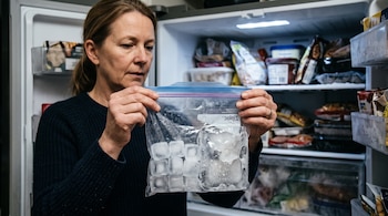Una mujer sostiene una bolsa plástica transparente con cubos de hielo separados y un bloque de hielo frente a un congelador abierto.
