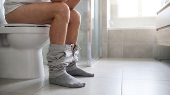 Un hombre con pantalones bajados hasta los tobillos y calcetines grises se sienta en un inodoro blanco dentro de un baño con baldosas claras.