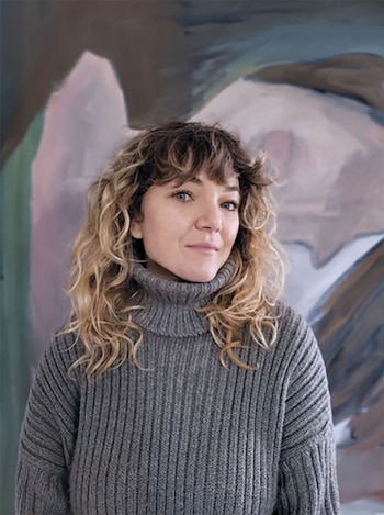 Mujer con cabello rizado rubio y flequillo, ojos azules, viste un suéter gris de cuello alto, de pie frente a una pintura abstracta