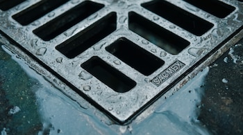 Fotografía de primer plano de una rejilla de coladera metálica con patrón de barras rectangulares y gotas de agua. Incluye el logotipo SACMEX.