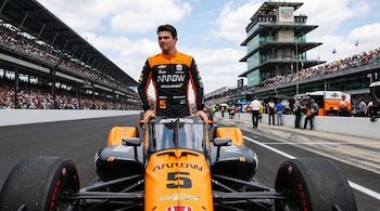 Pato O'Ward, vestido con un mono naranja y negro, de pie detrás de su coche de carreras naranja y negro número 5 en la pista de la Indy 500, con tribunas y una torre de control al fondo.