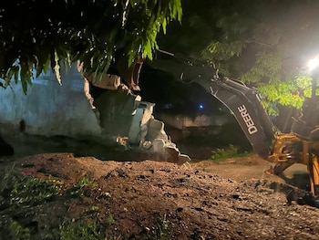 Vista nocturna de una excavadora John Deere amarilla y negra derribando una pared de concreto. Escombros en el suelo, hojas de árboles verdes arriba