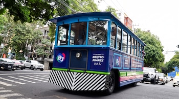 Autobús azul con diseño de tranvía rotulado con el texto de la alcaldía Benito Juárez que circula por una calle asfaltada entre vehículos.