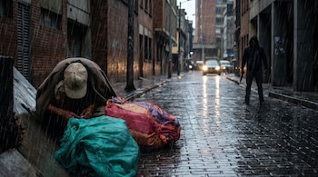 Vista en picado de un indigente cubierto con una manta en una calle mojada de Bogotá bajo la lluvia, con una figura con capucha y dos armas en la distancia.