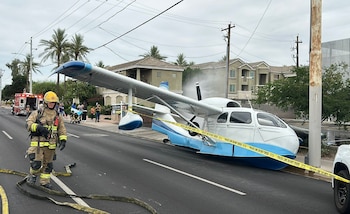 Un avión anfibio blanco y azul en una calle bloqueada por cinta policial, con un bombero en primer plano, una ambulancia y edificios residenciales al fondo