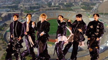 Seis miembros del grupo BTS vistiendo ropa negra posan delante del fondo de un estadio de fútbol y su entorno urbano.
