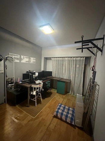 Interior de una habitación con paredes grises y suelo de madera. Se observa un escritorio con monitores, una silla, barra de dominadas, radiador y almohada