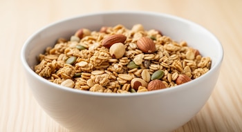 Primer plano de un cuenco de cerámica blanca con granola, almendras, avellanas y semillas de calabaza, sobre una mesa de madera clara.