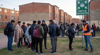 Grupo de personas de pie en área verde frente a edificios de ladrillo agrietados. Hombre reparte botellas de agua junto a cartel Punto de Evacuación.