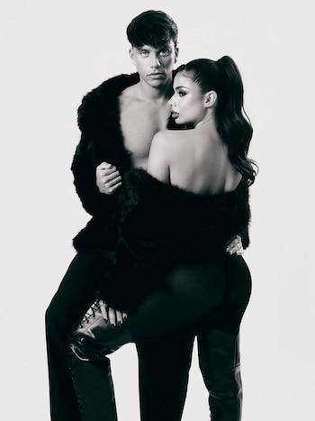 Foto en blanco y negro de Daniela Celis y Nick Sicaro. Él sin camisa con abrigo de piel, ella de espaldas con el mismo abrigo, cola de caballo y pierna levantada