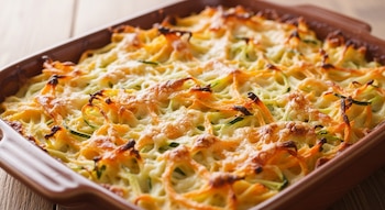 Imagen de un gratinado de zanahoria y calabacín en espiral con queso dorado y burbujeante, servido en una fuente de cerámica rectangular marrón sobre madera.