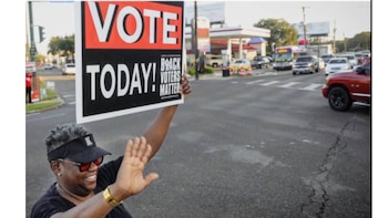 Una persona con gorra y gafas de sol sonríe mientras alza un cartel blanco y negro que dice "VOTE TODAY!" en una calle con tráfico de vehículos