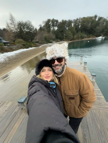 Un hombre y una mujer sonríen a la cámara. Ambos visten abrigos y gorros de invierno. Están en un muelle de madera junto a un lago con árboles y nieve en la orilla