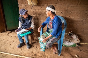 Dos mujeres conversan sentadas en una habitación con pared de adobe, una con chaleco azul y gorra escribiendo en un portapapeles