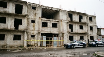 Un edificio de concreto sin terminar con varillas expuestas, cinta amarilla de no pasar y dos patrullas policiales en la calle.