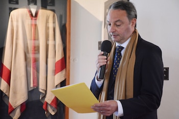 Un hombre con traje, bufanda beige y corbata habla por un micrófono mientras sostiene un documento amarillo junto a un poncho en exhibición
