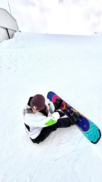 Una mujer con gorro y abrigo de invierno permanece sentada en la nieve junto a una tabla de snowboard