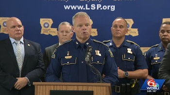 Um policial de uniforme azul fala em um pódio com um microfone, junto com outros policiais e homens uniformizados.