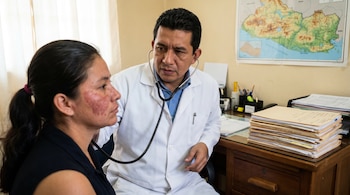 Un médico varón con bata blanca examina a una mujer con sarpullido rojizo en el rostro usando un estetoscopio en una consulta médica.