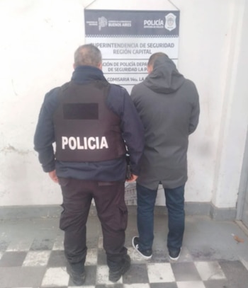 Un agente de policía con chaleco negro y la palabra "POLICIA" en blanco, junto a un hombre vestido de gris y jeans, de espalda, frente a un cartel