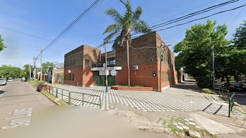 Fachada de un edificio de ladrillo rojo con rejas verdes, una palmera y cables eléctricos. Hay una calle y una vereda con baldosas a cuadros