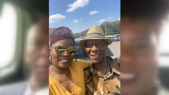 Un hombre y una mujer sonríen a la cámara al aire libre. El hombre usa sombrero, la mujer gafas de sol. Hay vehículos y árboles en el fondo