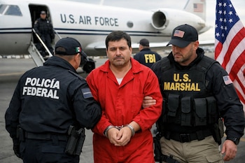 Un hombre esposado con mono naranja es escoltado por dos agentes con uniformes de Policía Federal y U.S. Marshal, frente a un avión de la Fuerza Aérea de EE. UU. y una bandera estadounidense.