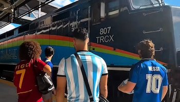 Vista trasera de personas con camisetas de fútbol, una de ellas con "MESSI 10", observando un tren oscuro con franjas de colores y el número 807 TRCX en una estación