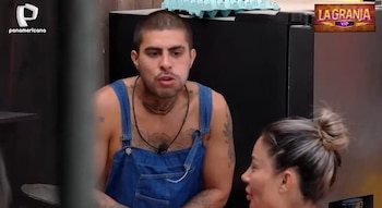 Paul Michael, con overol de jean y tatuajes, habla intensamente a una mujer con cabello recogido, en un entorno interior con logos de 'Panamericana' y 'La Granja VIP'
