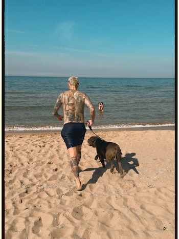 Hombre tatuado de espaldas, con pantalones cortos, corre descalzo en la arena de la playa con un perro oscuro con correa; el mar azul al fondo