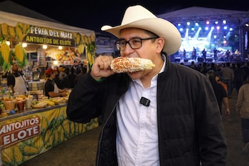 Un hombre con sombrero vaquero y gafas come un elote. Detrás, un puesto de comida "Feria del Maíz Antojitos de Elote" y un escenario con luces y banda musical.