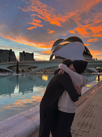 Vista trasera de una pareja abrazándose junto a un estanque. Al fondo, edificios modernos y un puente bajo un espectacular atardecer anaranjado
