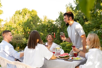 Cinco jóvenes adultos comparten una comida y bebidas en una mesa al aire libre, con vasos de líquido rojo y sandía, sonriendo y riendo bajo el sol