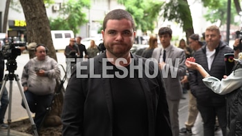 Un hombre caucásico de barba y chaqueta negra mira al frente, rodeado de cámaras de televisión, trípodes, y varias personas en una calle arbolada