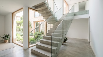 Escalera de hormigón con barandilla de vidrio en un interior de paredes blancas, suelo pulido y ventanal con vista a un jardín.