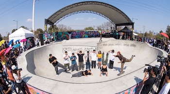 Jóvenes en distintas posturas dentro de una pista de patinaje de concreto, con espectadores, cámaras y sombrillas alrededor.