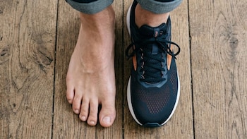 Primer plano de dos pies sobre suelo de madera: uno descalzo y otro con zapatilla de running oscura con detalles naranjas y cordones negros