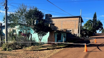 Vista exterior de una propiedad con cinta de precaución y conos naranjas en un camino de tierra. Una casa azul y otra de ladrillo bajo un cielo claro