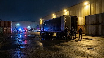 Un camión con el remolque abierto, agentes con chalecos tácticos, contenedores y patrullas con luces encendidas en un playón nocturno y mojado.