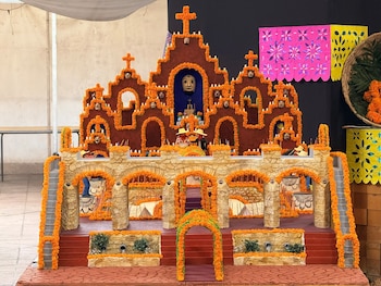 Maqueta de una iglesia decorada con flores naranjas de cempasúchil, pequeñas veladoras, figuras y papel picado rosa al fondo