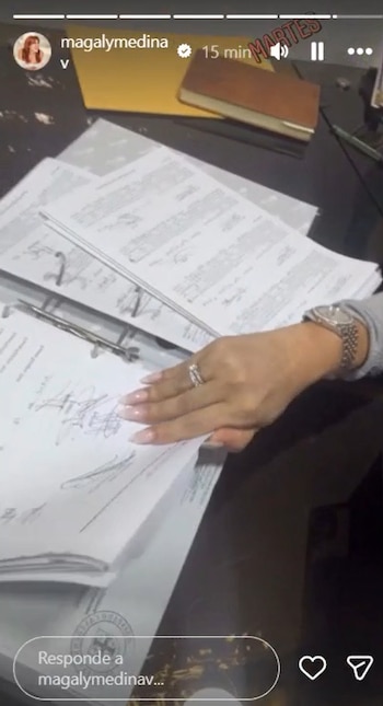 Una mano de mujer con anillo y reloj de plata hojea un cuaderno de anillas con documentos sobre un escritorio oscuro, junto a una carpeta amarilla y un cuaderno marrón