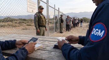 Manos revisan pasaportes y documentos sobre mesa de madera. Agentes uniformados de migración dominicana y personas en fila junto a una valla metálica.