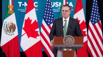 Un hombre con traje oscuro y corbata verde habla en un podio con el escudo de México. Detrás, banderas de México, Canadá y Estados Unidos, junto a un letrero de T-MEC/USMCA.